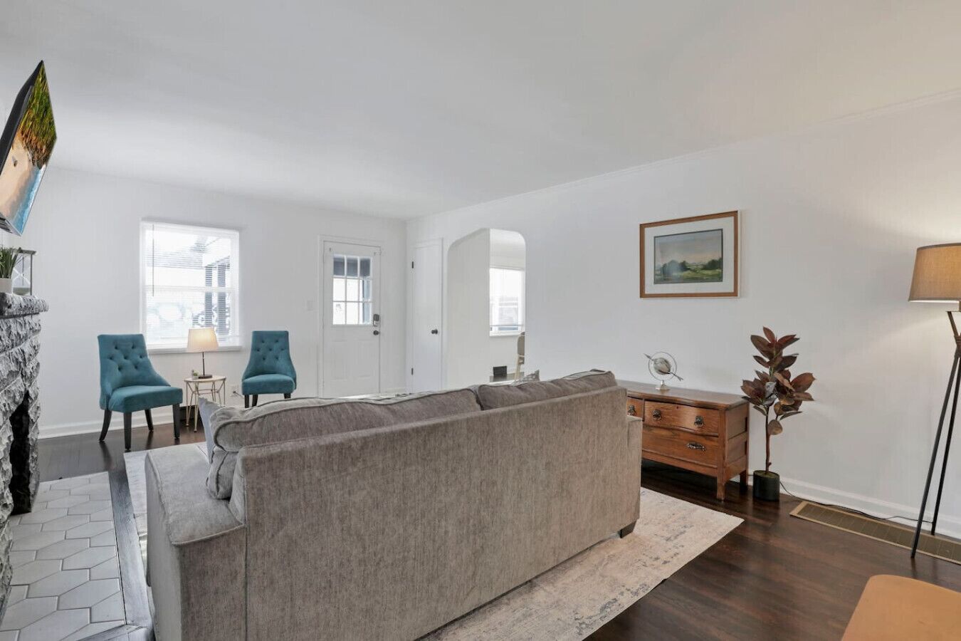 Een woonkamer met een grijze bank, twee blauwe fauteuils, een houten dressoir en een vloerlamp.