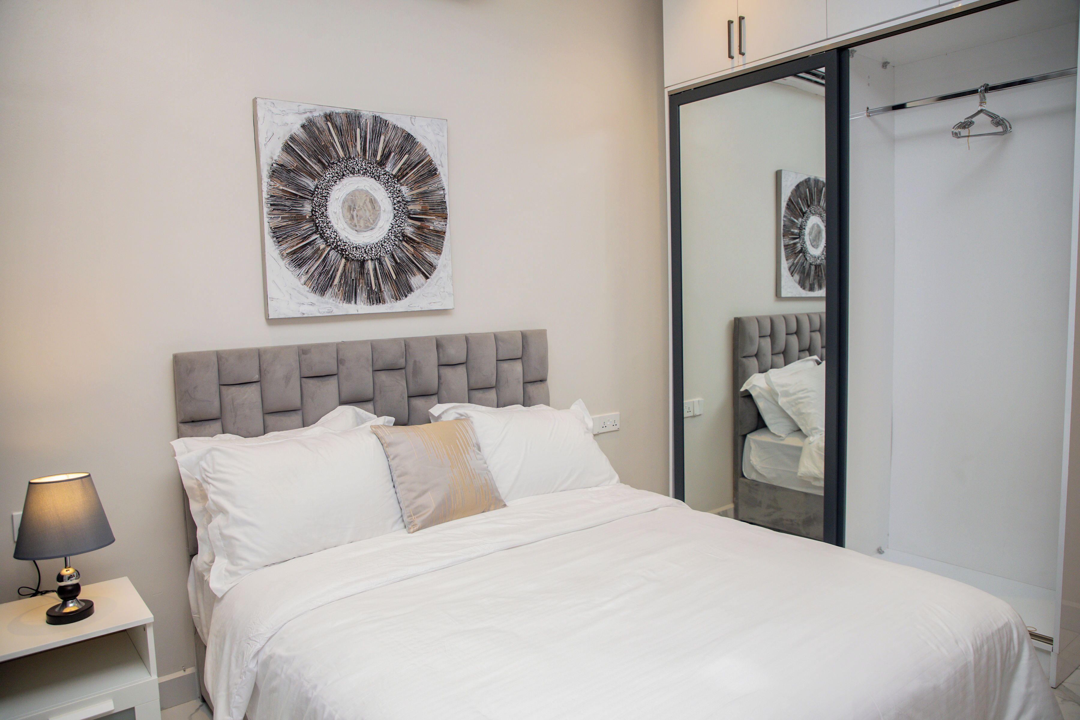 A bedroom with a bed, a bedside table with a lamp, a large mirror, and a wall-mounted artwork.