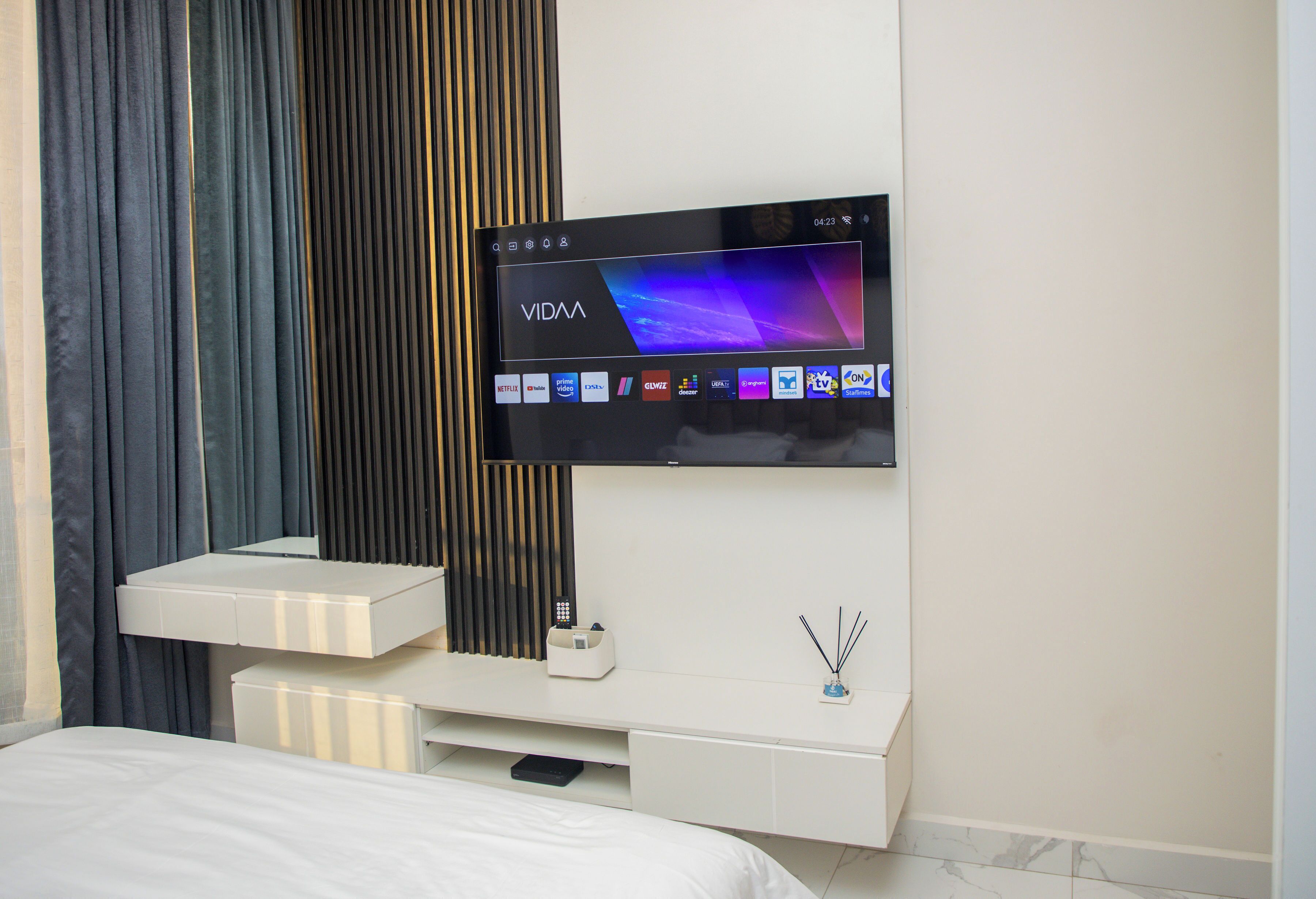 A modern hotel room with a flat-screen TV mounted on the wall, a white bedside table, and a bed with white bedding.