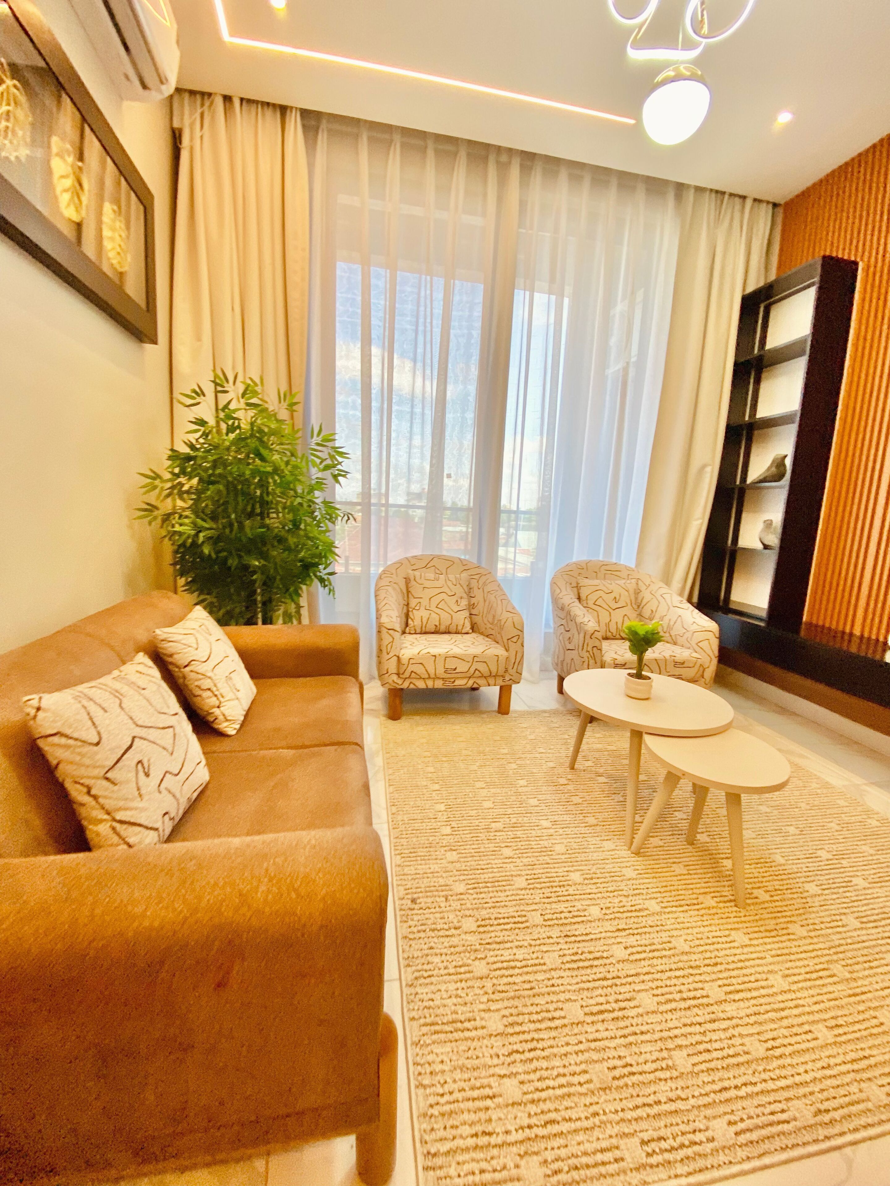 Ein Wohnzimmer mit einem braunen Sofa, gemusterten Sesseln, einem kleinen runden Tisch und einem großen Fenster mit durchsichtigen Vorhängen.