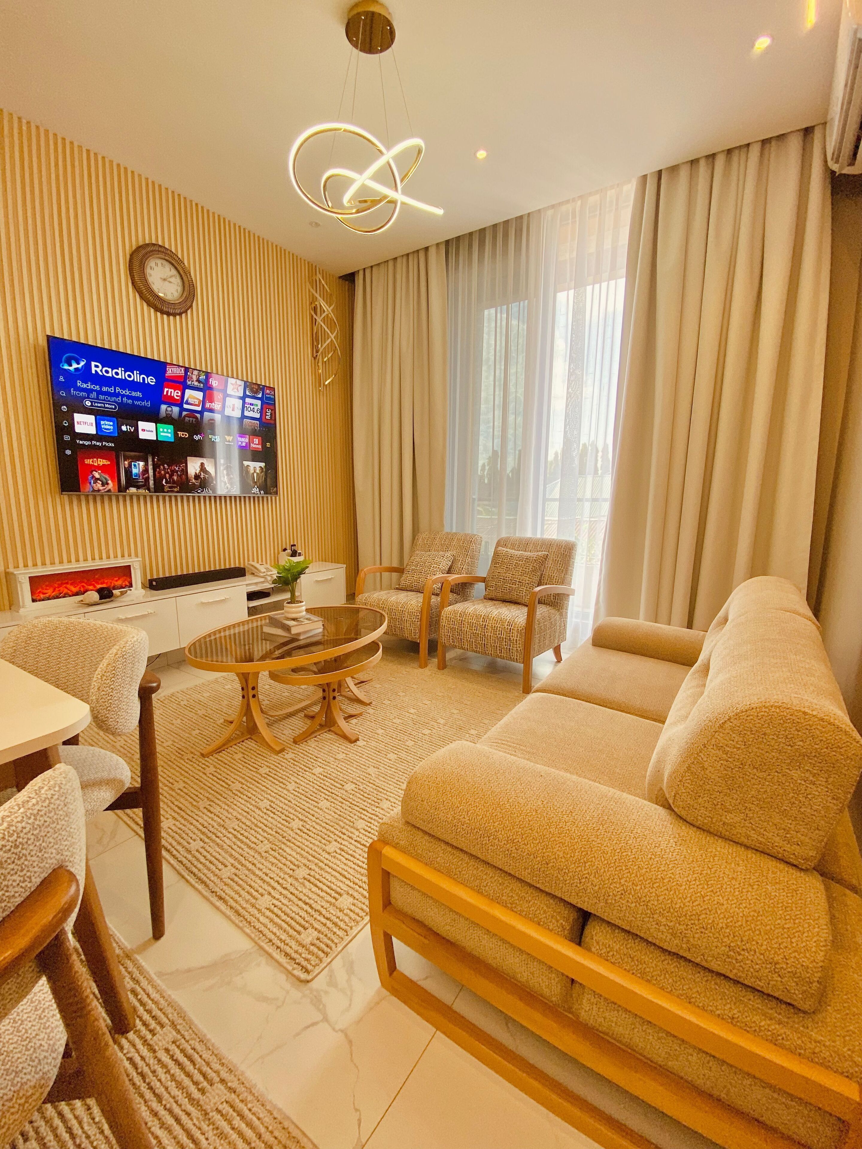 Una sala de estar moderna con un televisor de pantalla plana mostrando varios canales, una mesa de centro redonda de vidrio, dos sillones estampados, un sofá color beige y una pared a rayas.