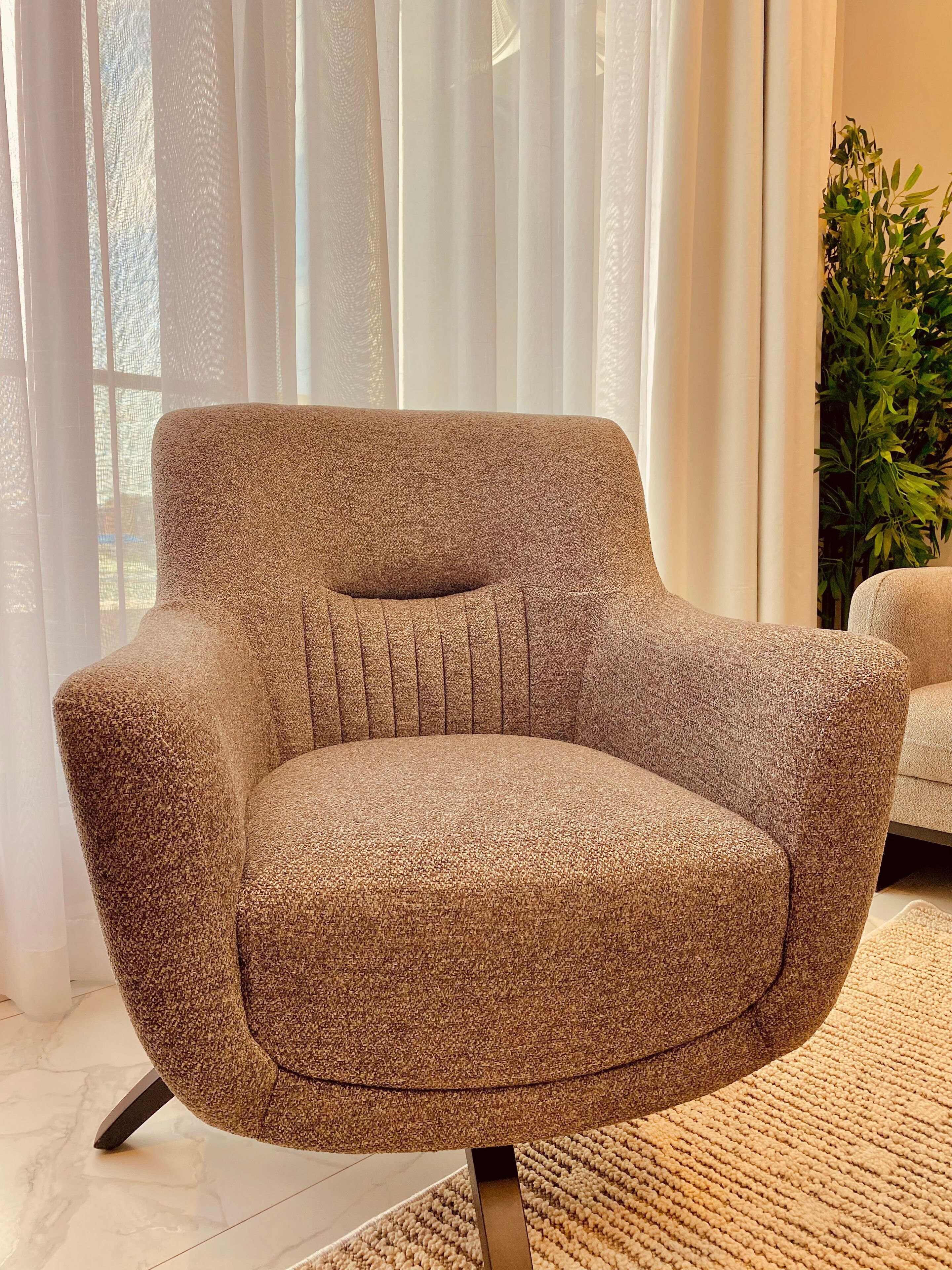 A modern armchair with a textured fabric, positioned in a well-lit room with sheer curtains and a potted plant.