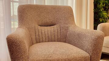 A modern armchair with a textured fabric, positioned in a well-lit room with sheer curtains and a potted plant.