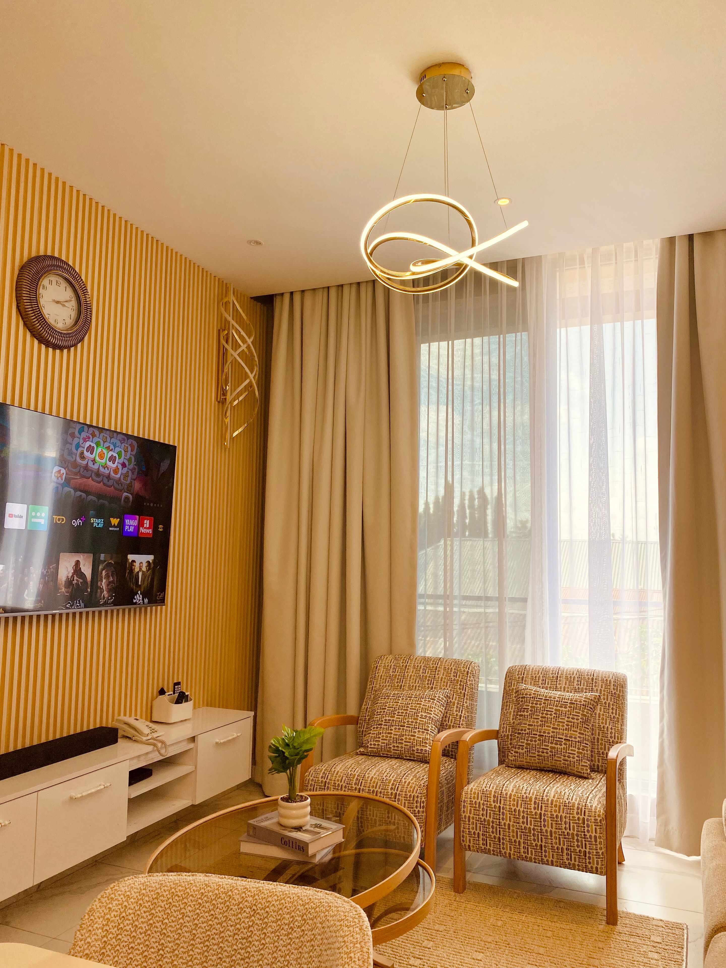 A modern living room with a flat-screen TV, two patterned armchairs, a coffee table, and a decorative light fixture.