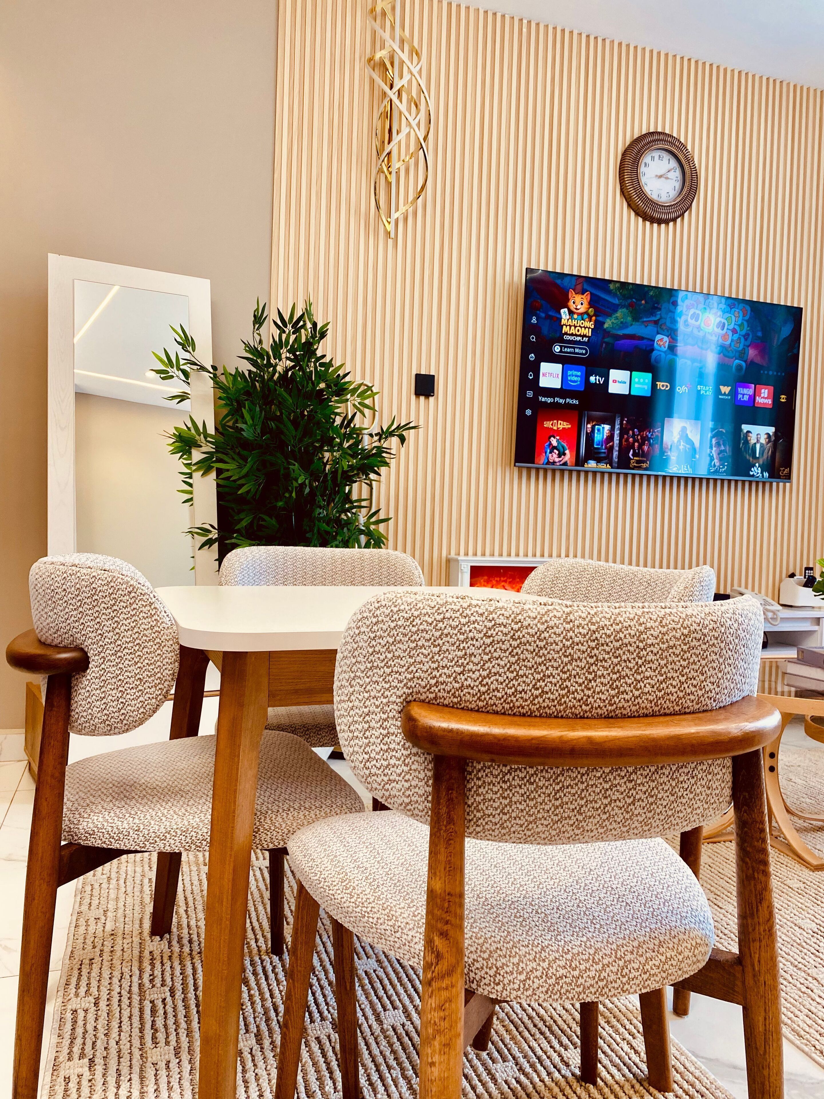A dining area with a round table, four upholstered chairs, a potted plant, a wall-mounted TV displaying various channels, and a clock.