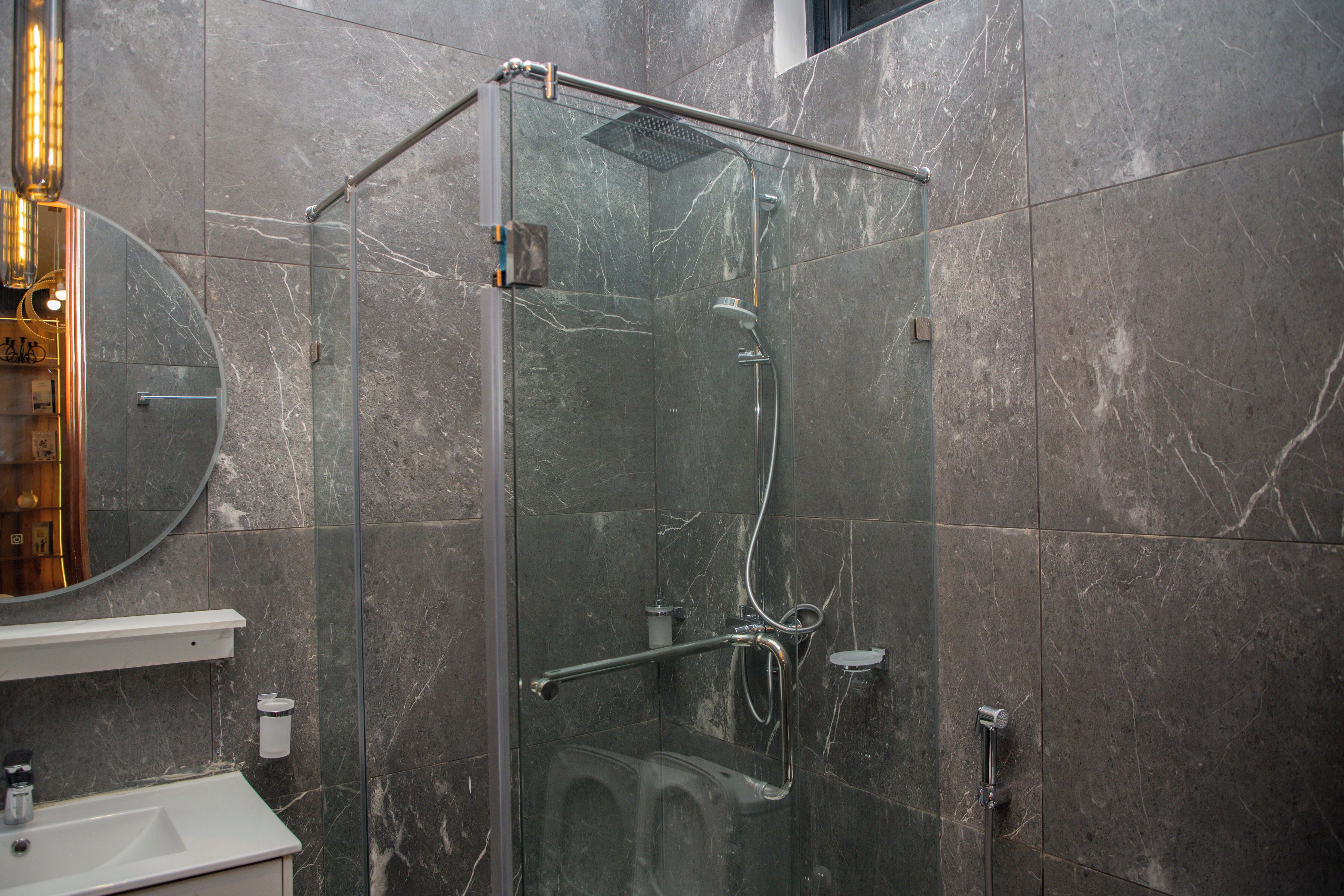 Un baño moderno con ducha envuelta en vidrio, pared con azulejos de mármol y lavamanos blanco con espejo arriba.