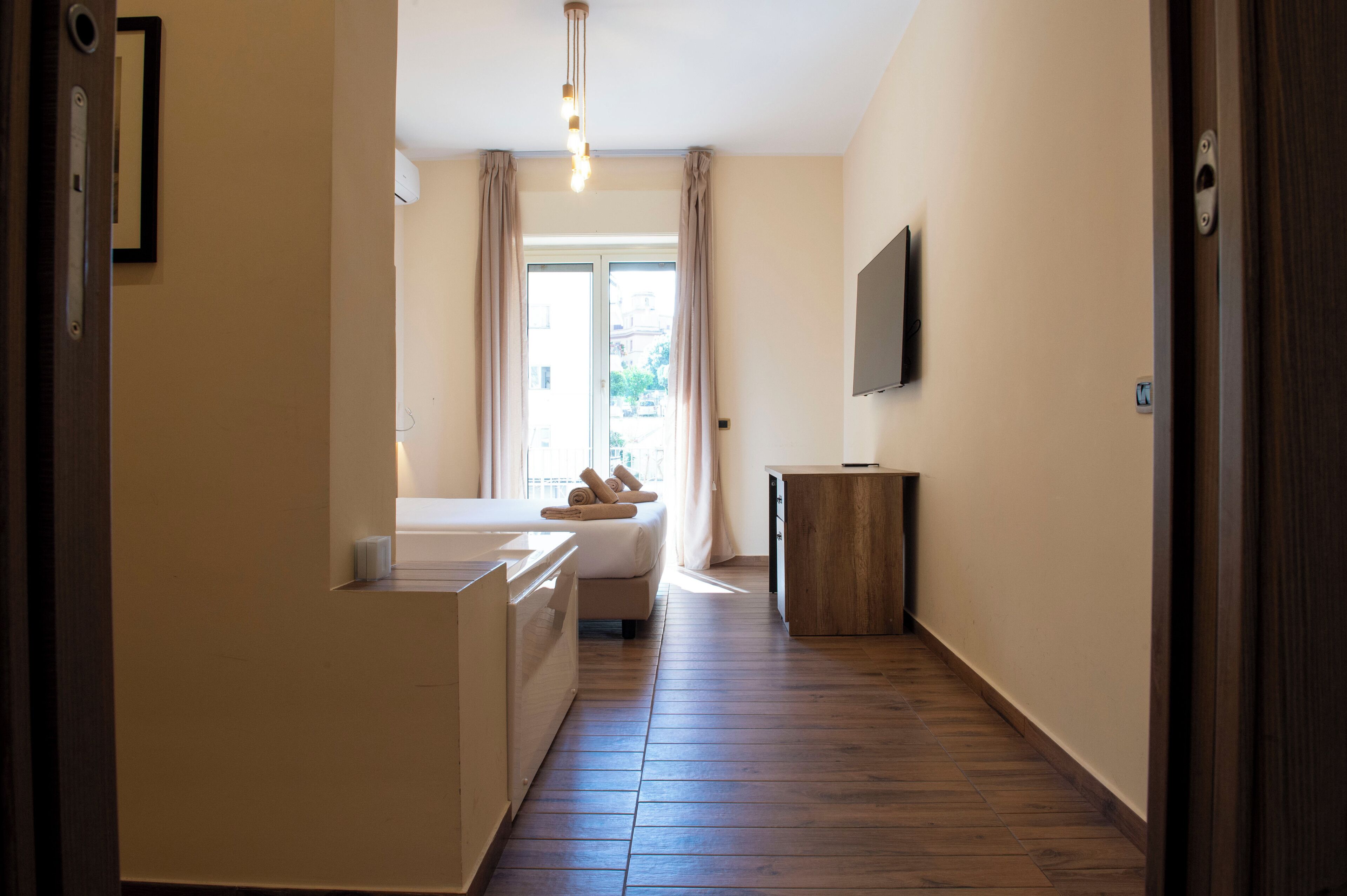 Una habitación de hotel con cama, una mesita de noche pequeña, suelo de madera, ventana con cortinas y un televisor montado en la pared.