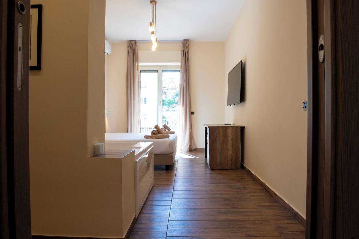 A hotel room with a bed, a small bedside table, a wooden floor, a window with curtains, and a television mounted on the wall.