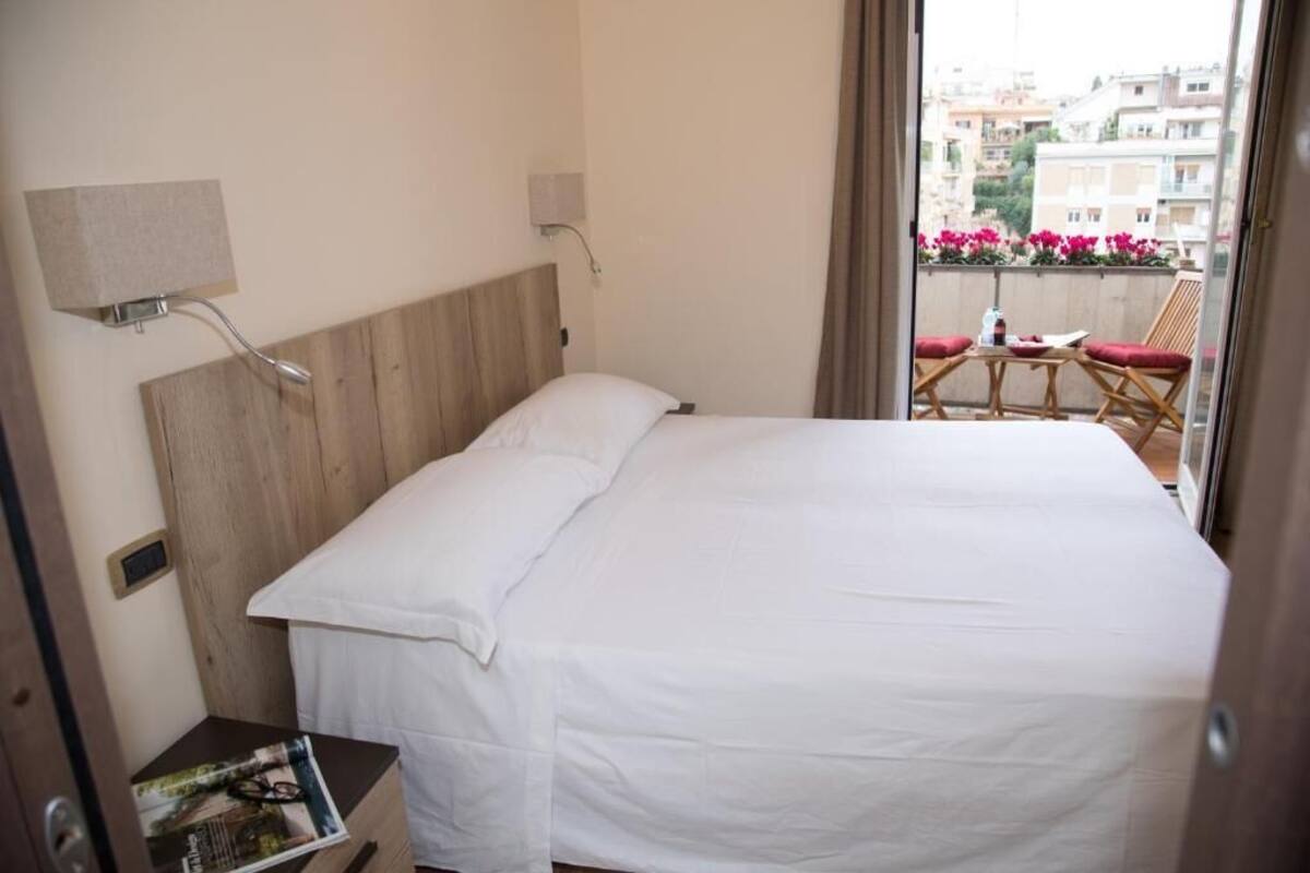 A hotel room with a wooden headboard, a bed with white linens, a small bedside table with a magazine, and a view of a balcony with red flowers and a table.