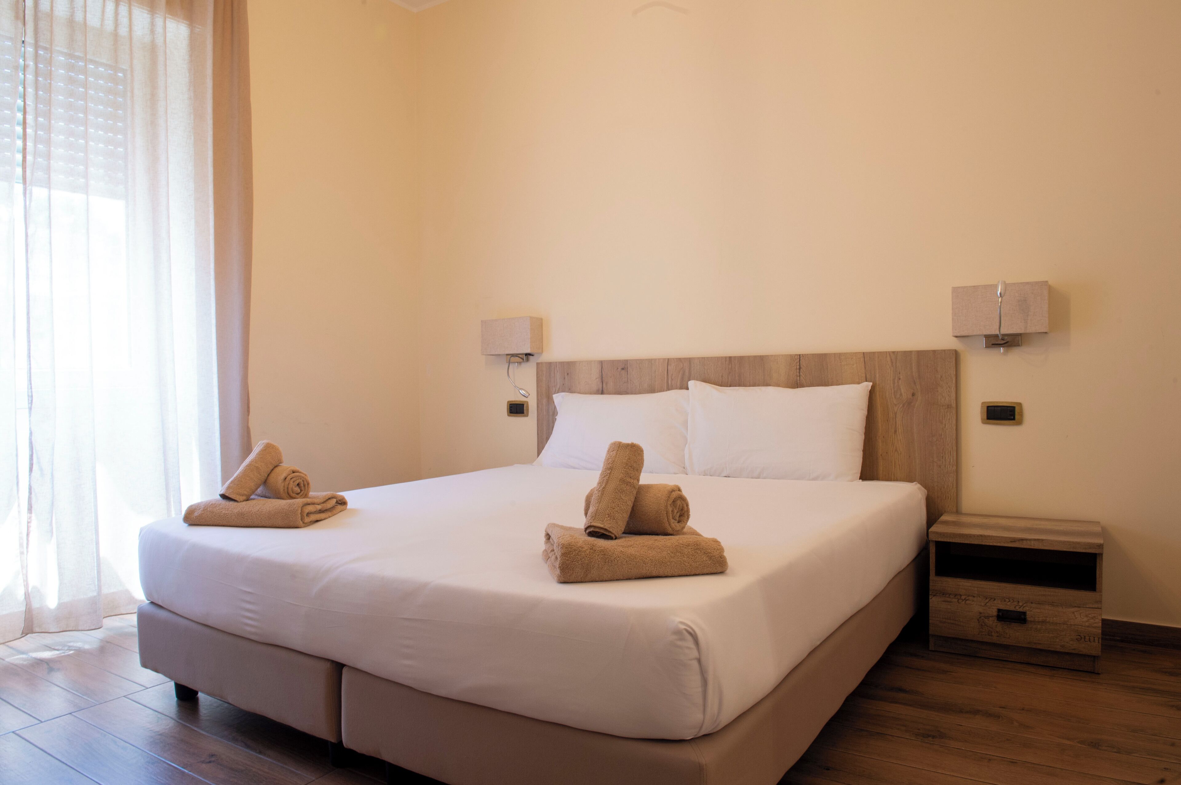 A neatly made bed with white linens and two rolled towels on the side. There are two wall-mounted nightstands with lamps.