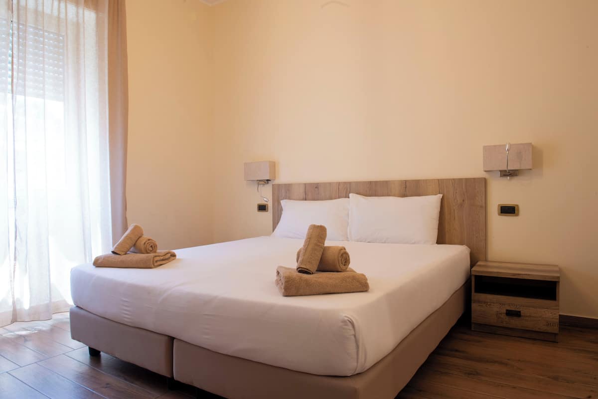 A neatly made bed with white linens and two rolled towels on the side. There are two wall-mounted nightstands with lamps.