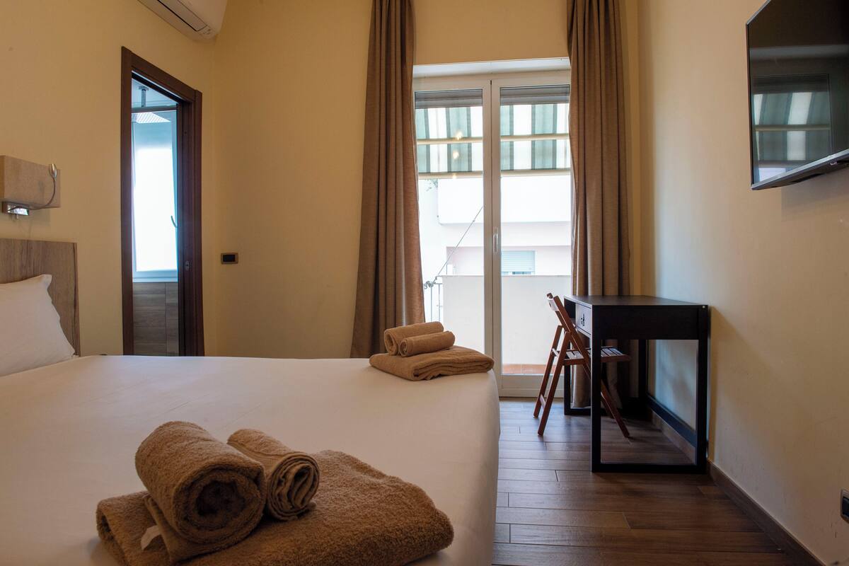 A hotel room with a bed, a desk, a TV, and a balcony with curtains.