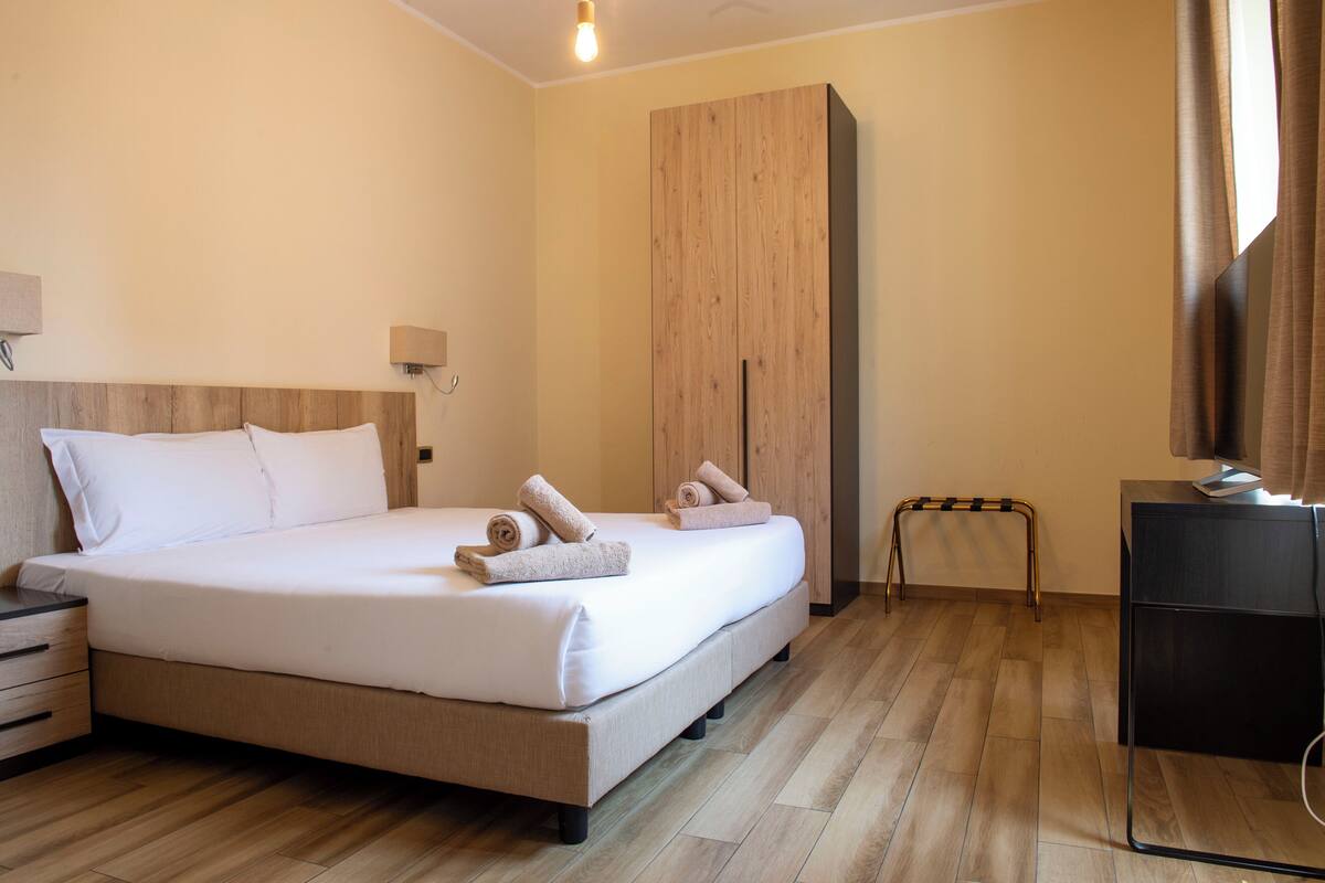 A neatly made bed with towels, a wooden headboard, a nightstand, a small table with a lamp, and a wardrobe.