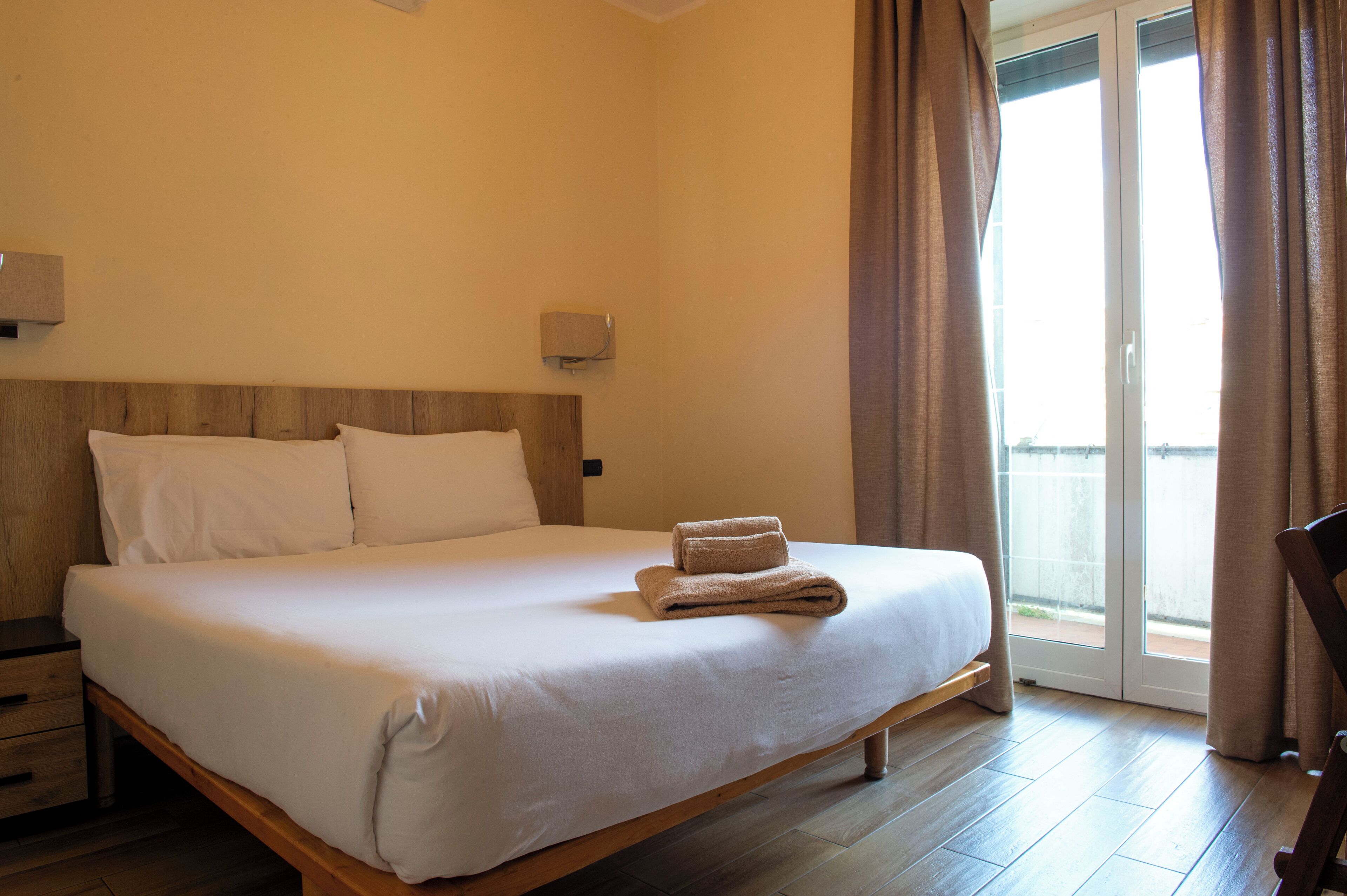 A hotel room with a large bed, wooden headboard, two bedside tables, a nightstand, and a sliding glass door leading to a balcony.