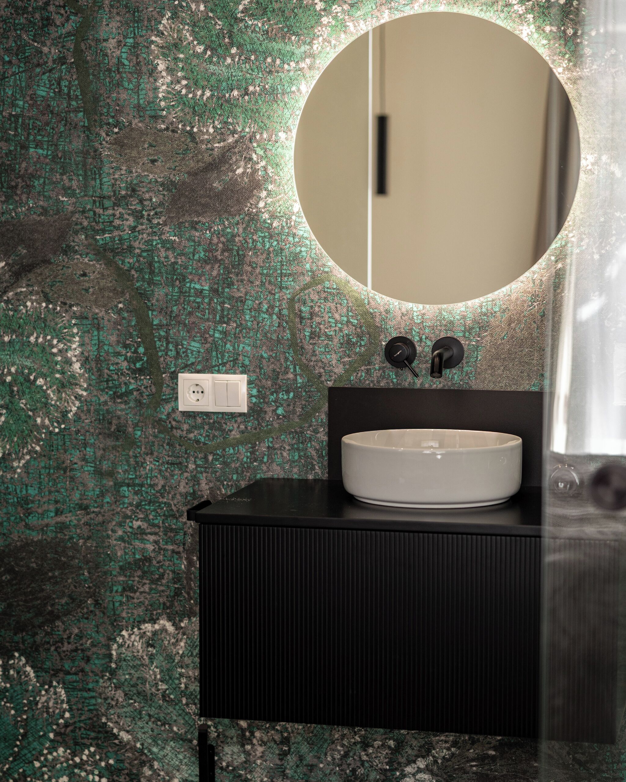 Een badkamer met een ronde spiegel, een witte wastafel en een wand met groene textuur.