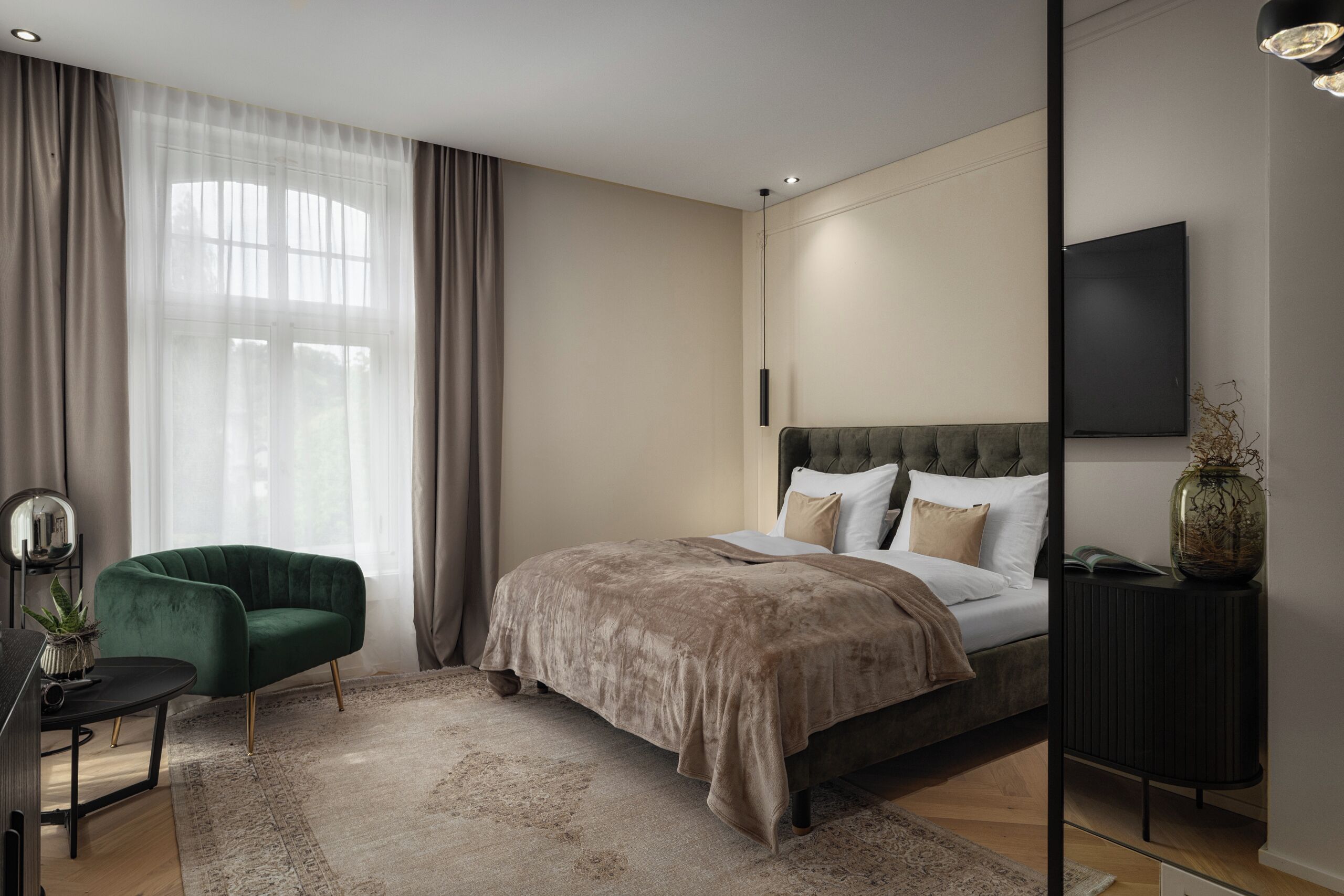 Un dormitorio moderno con una cama grande, un sillón verde, una mesita de noche con un florero y un televisor en la pared.