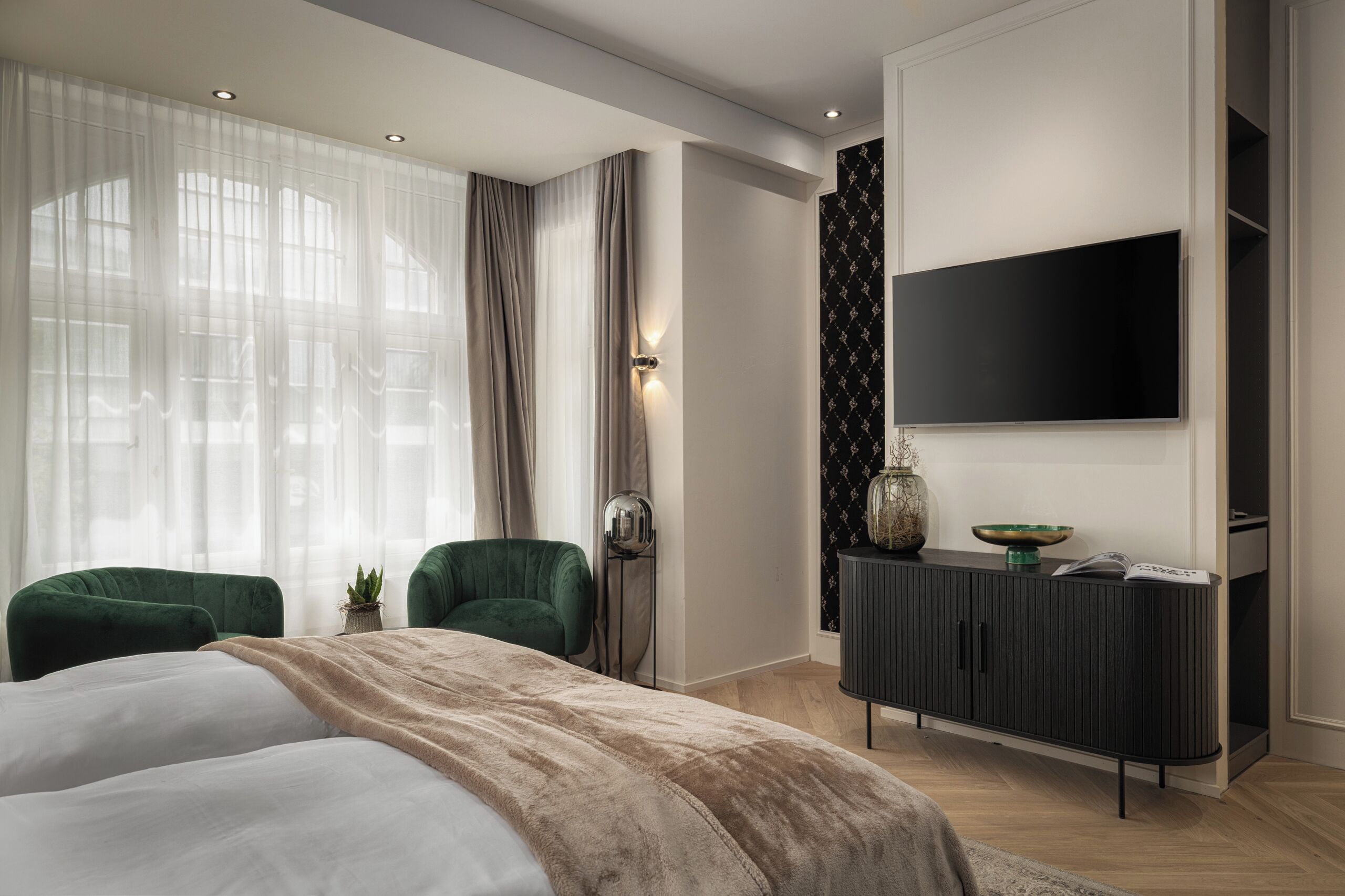 A modern bedroom with a large bed, two green armchairs, a black TV on the wall, and a dark wooden cabinet.