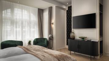 Une chambre moderne avec un grand lit, deux fauteuils verts, un téléviseur noir fixé au mur et une commode en bois foncé.
