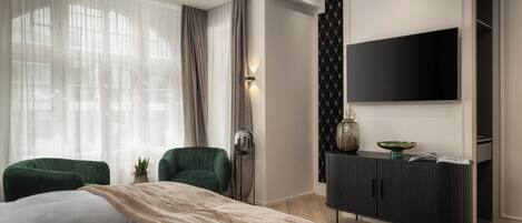 A modern bedroom with a large bed, two green armchairs, a black TV on the wall, and a dark wooden cabinet.