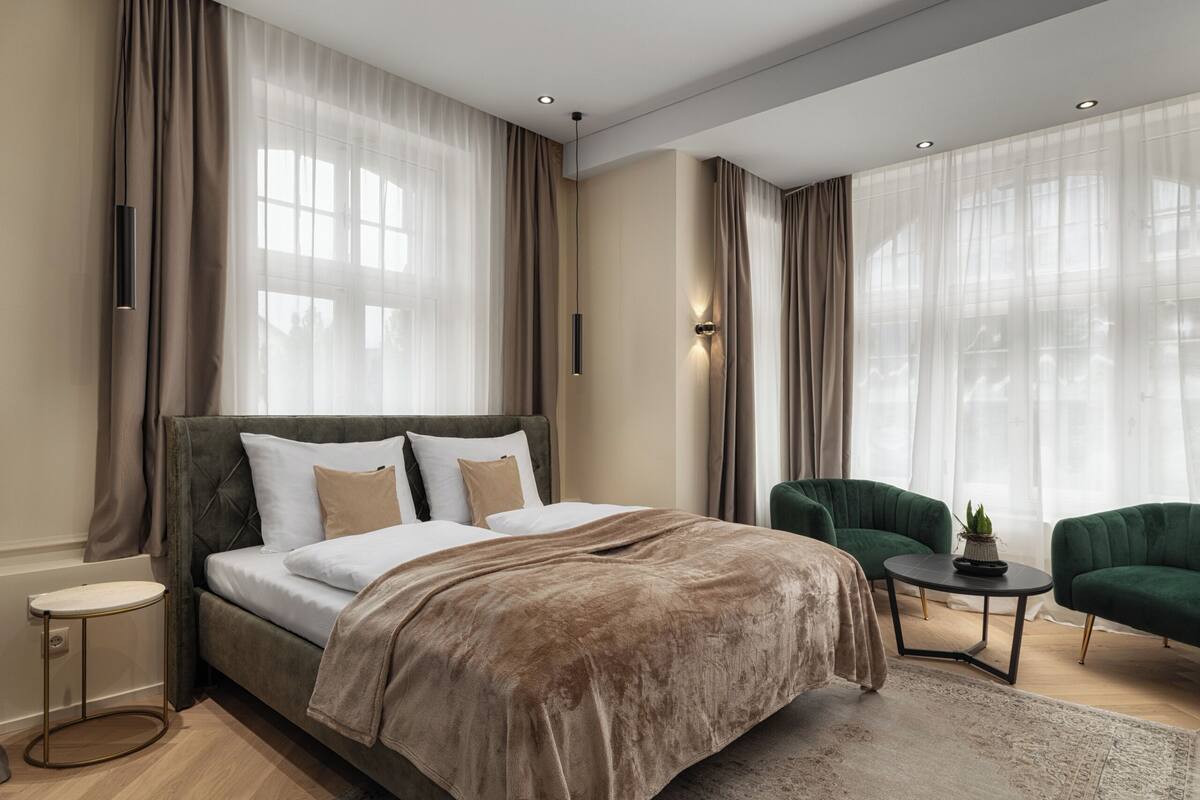 A modern bedroom with a large bed, two green armchairs, a small round table, and sheer curtains.