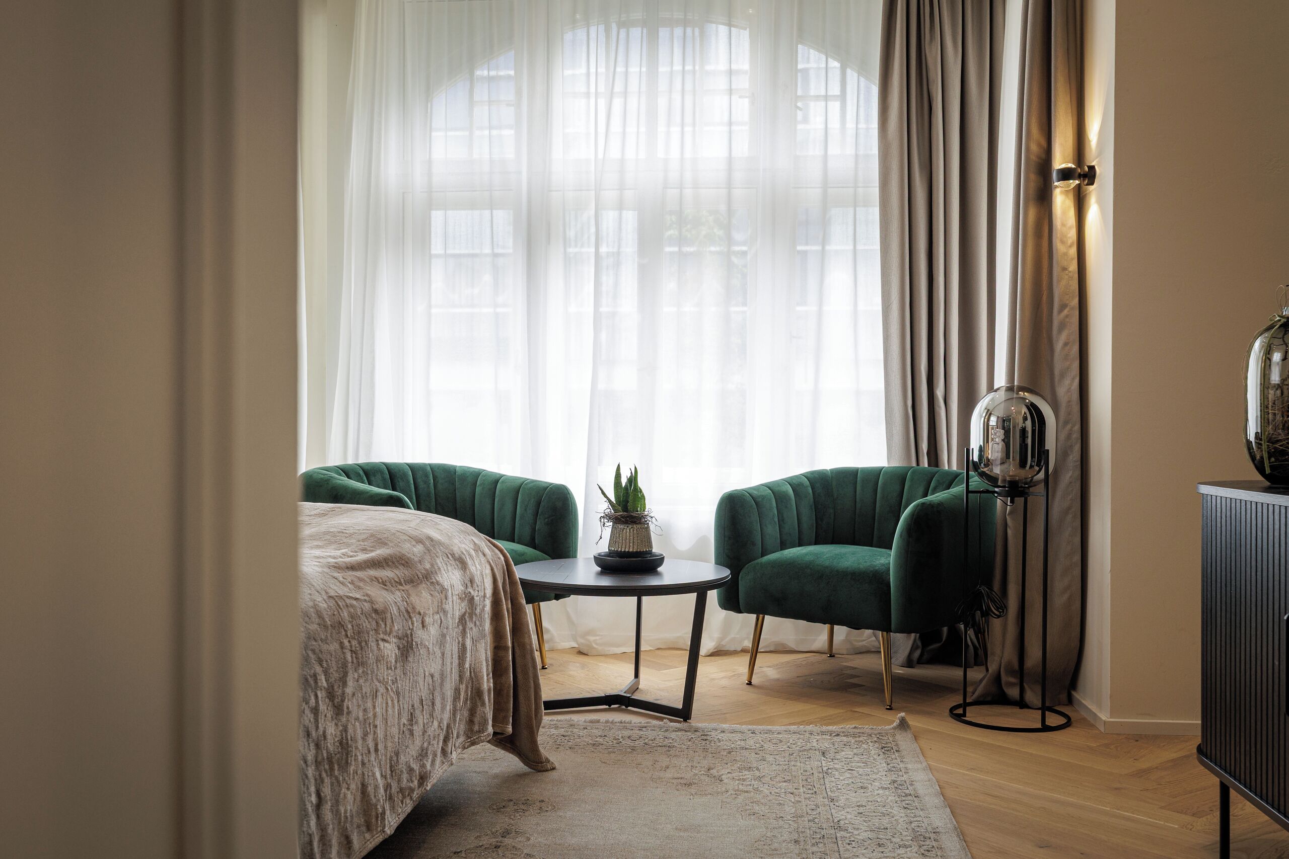 A bedroom with a bed, two green armchairs, a small table with a plant, and a floor lamp.