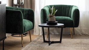 Una habitación con dos sillones verdes, una pequeña mesa redonda con una planta en maceta, un objeto esférico reflectante y un amplio ventanal con cortinas traslúcidas.