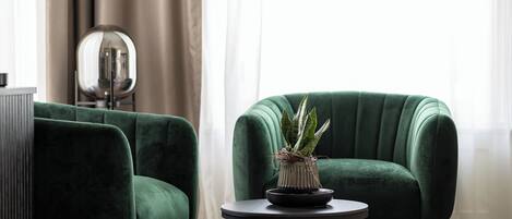 Une pièce avec deux fauteuils verts, une petite table ronde ornée d’un pot de fleurs, un objet sphérique réfléchissant et une grande fenêtre avec des rideaux légers.