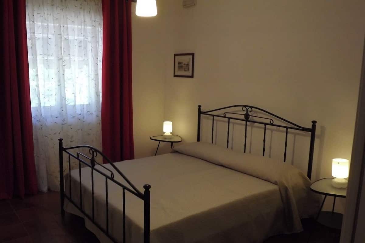 Une chambre avec un lit simple, deux tables de chevet, une fenêtre avec des rideaux rouges et un tableau encadré au mur.