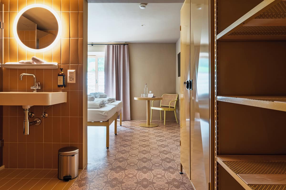 A modern hotel bathroom with a sink, mirror, and toilet. There is a small table with a chair and a bed in the background.