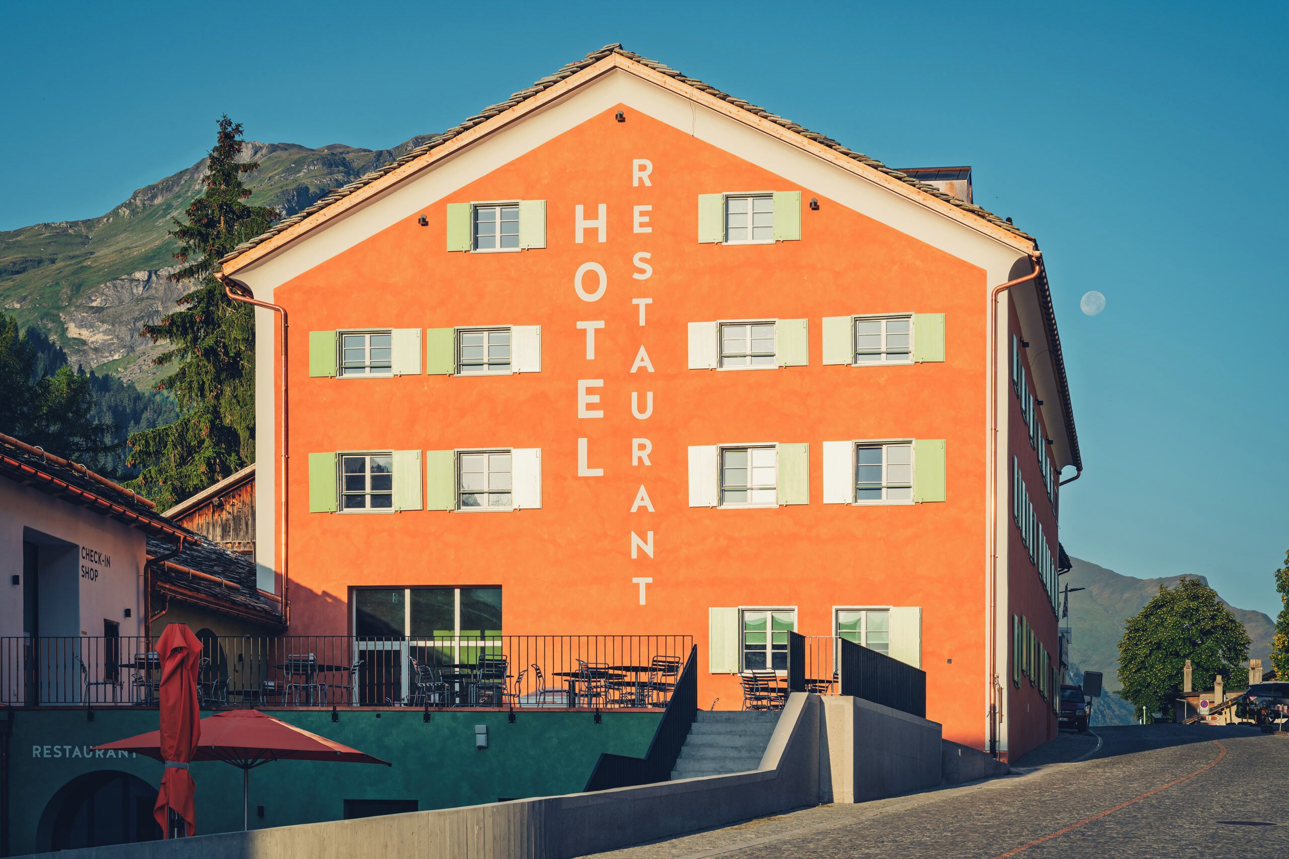 A multi-story building with the words "RESTAURANT HOTEL" on its facade, a staircase leading to the entrance, and outdoor seating with tables and chairs.