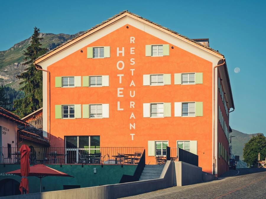 A multi-story building with the words "RESTAURANT HOTEL" on its facade, a staircase leading to the entrance, and outdoor seating with tables and chairs.