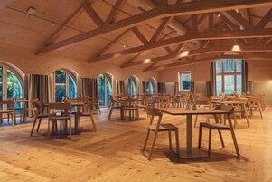 Un amplio salón con suelos de madera y mesas, grandes ventanales de arco y un alto techo de madera.