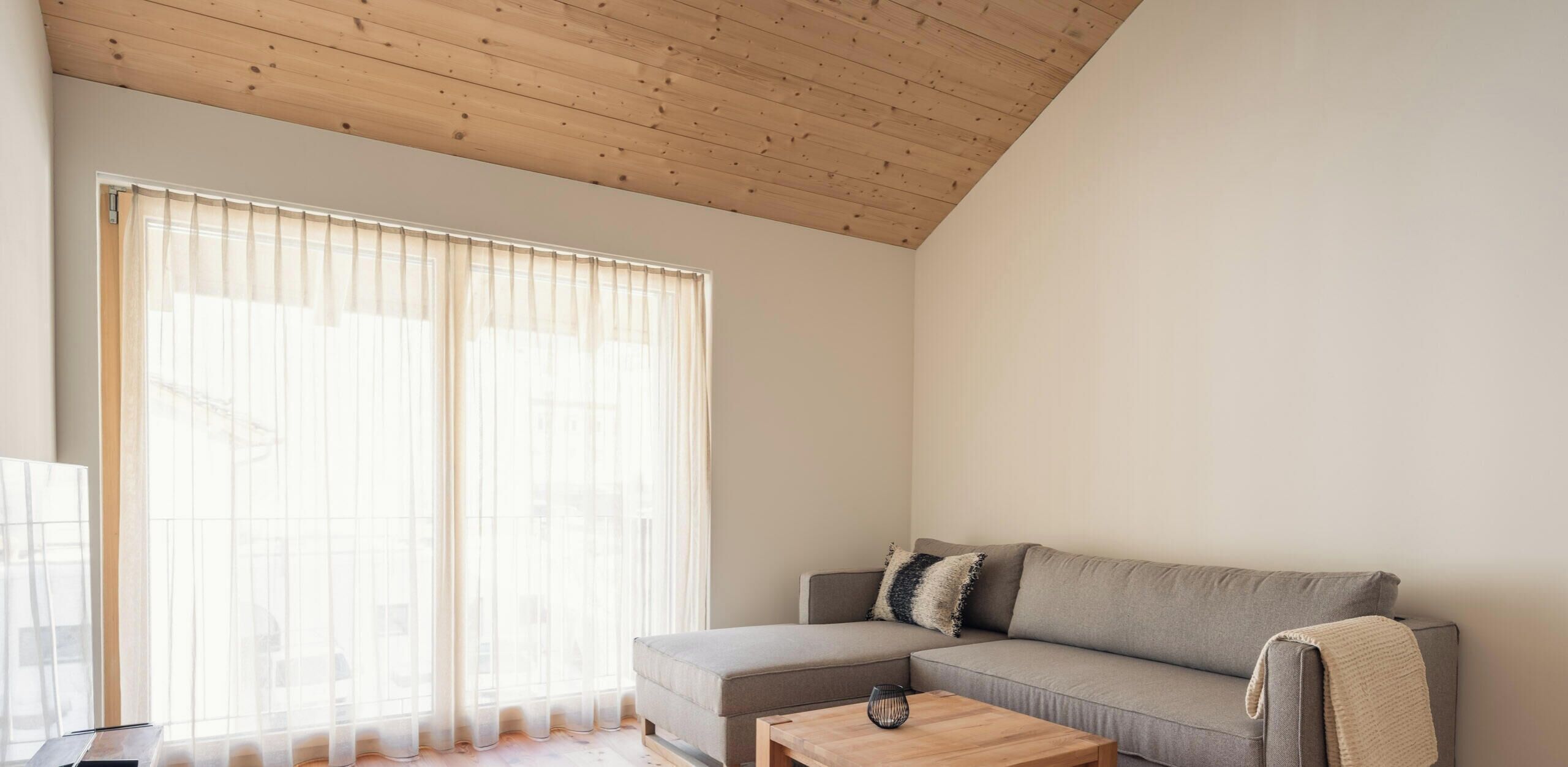 A modern living room with a wooden ceiling, a grey sofa, a small coffee table, and sheer curtains on a large window.