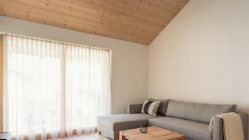 A modern living room with a wooden ceiling, a grey sofa, a small coffee table, and sheer curtains on a large window.