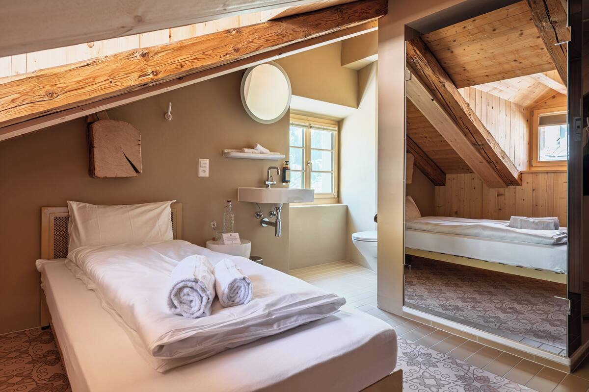 A bedroom with a bed, a bedside table, a mirror, a window, and a view of another room.