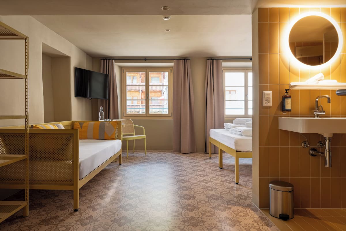 A hotel room with a bed, a sofa, a TV, a window with curtains, a bathroom with a sink and mirror, and a patterned floor.