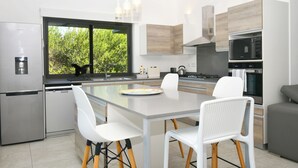 A modern kitchen with a central island, white chairs, stainless steel appliances, and a view of greenery outside.
