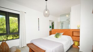 A bedroom with a large bed, a wooden headboard, a bench, a window with curtains, a hanging light fixture, and a mirror.