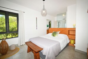 Ein Schlafzimmer mit einem großen Bett, einem hölzernen Kopfteil, einer Sitzbank, einem Fenster mit Vorhängen, einer Pendelleuchte und einem Spiegel.
