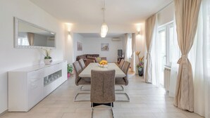 A modern dining area with a white table, brown chairs, and a chandelier. There is a white cabinet with drawers and a mirror on the left. The room has beige curtains and a large window on the right. - Evita Apartment - 60m to Beach, A/C in All Sleeping Areas! (Slatine)