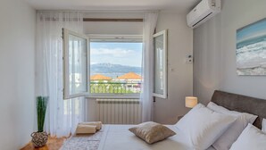 A bedroom with a bed, a window with curtains, a radiator, a wall-mounted air conditioner, and a view of buildings and mountains. - Evita Apartment - 60m to Beach, A/C in All Sleeping Areas! (Slatine)