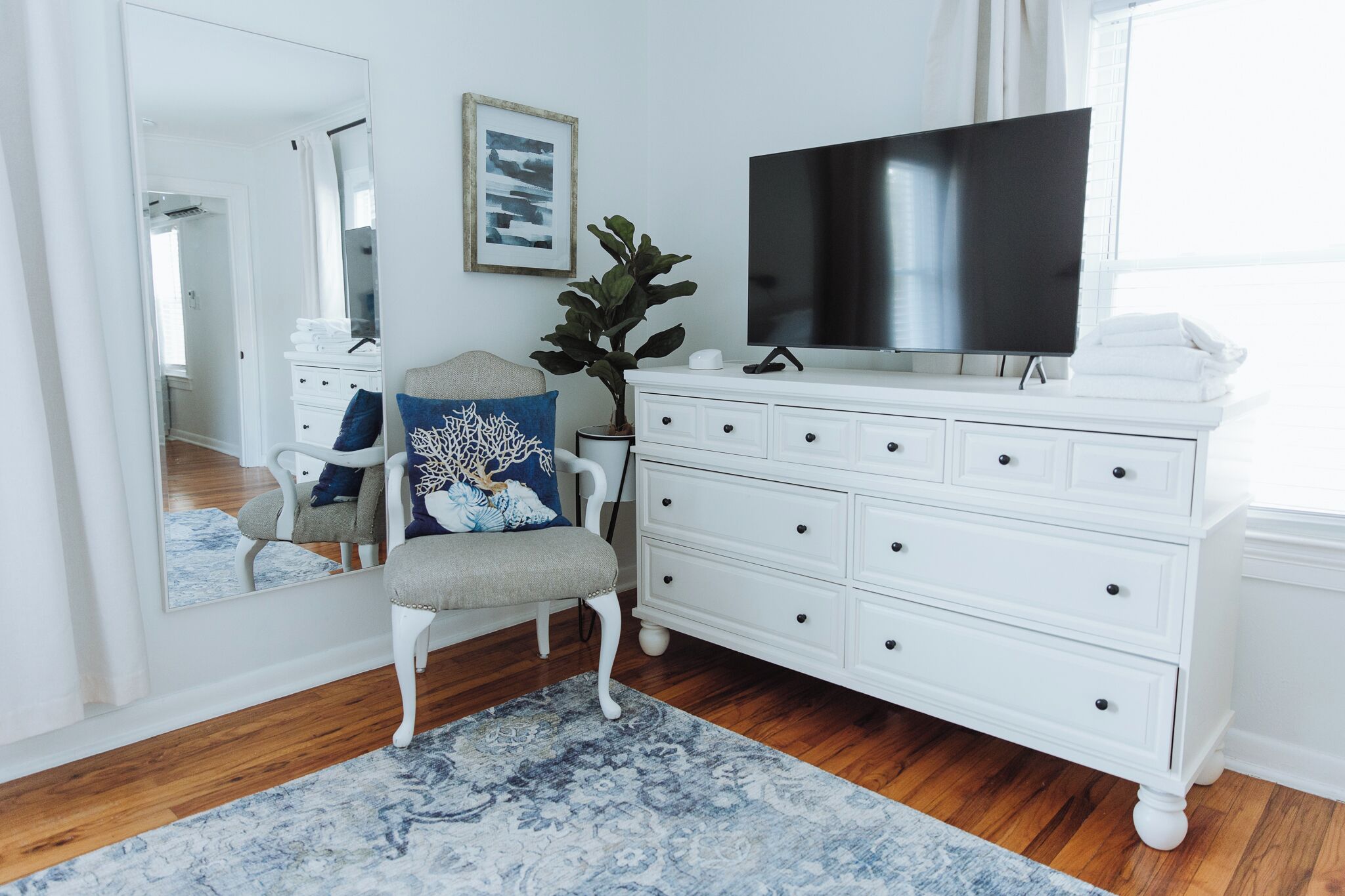 A bedroom with a white dresser, a flat-screen TV, a mirror, and a chair with a blue cushion.