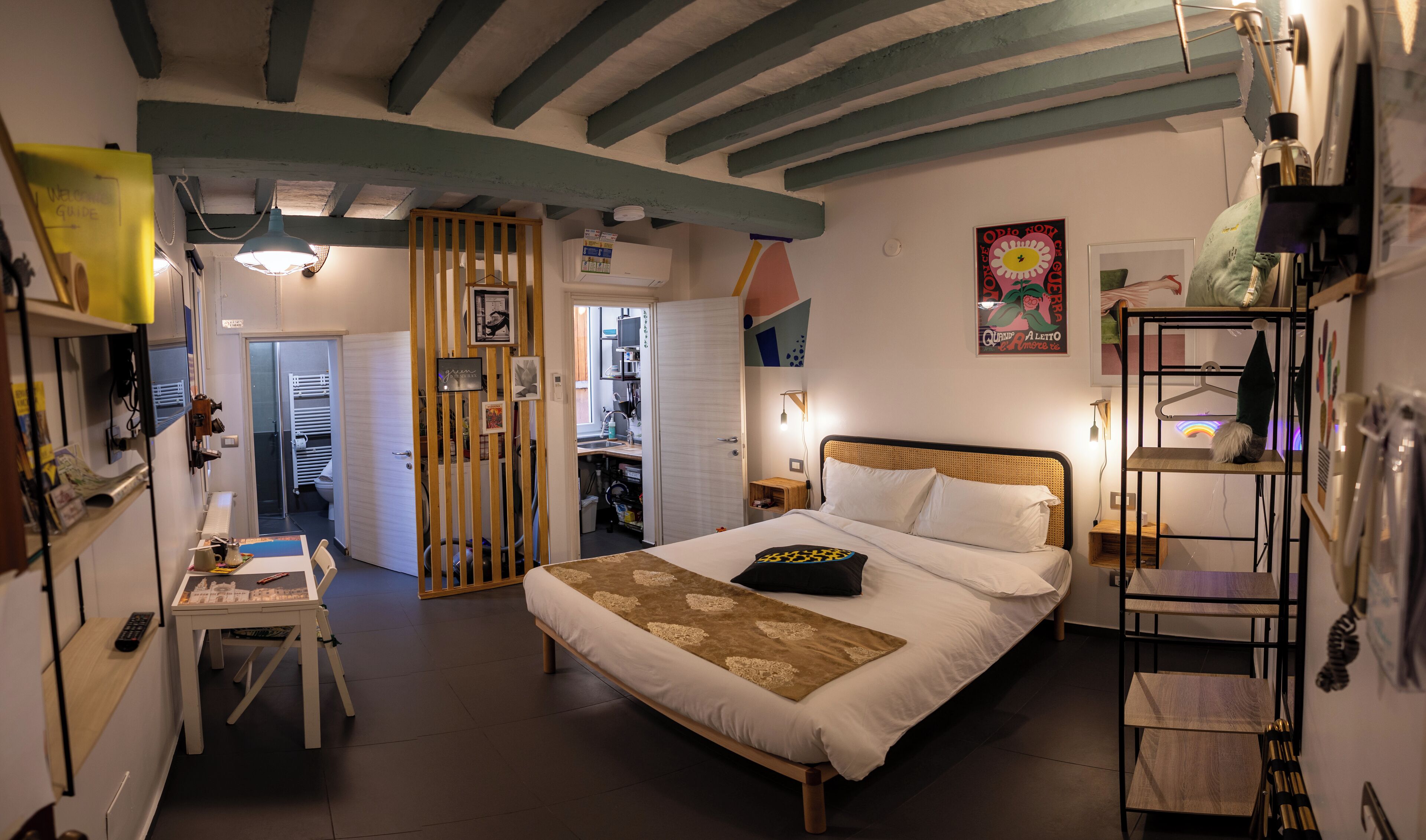 A bedroom with a bed, bedside table, and a desk with a chair. There is a wardrobe and a door leading to another room.
