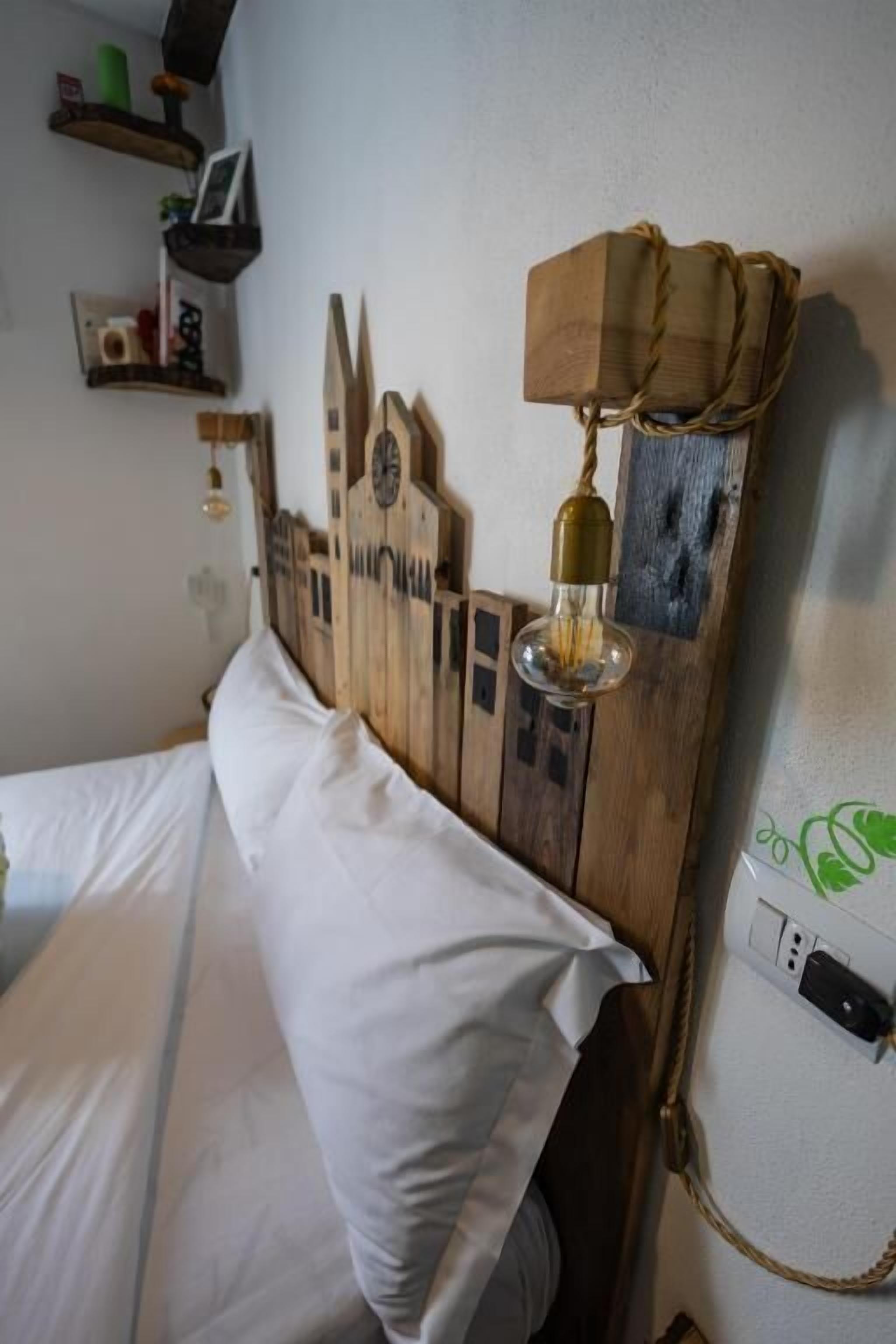 A bed with white linens, a wooden headboard with a clock, and a wall-mounted lamp.