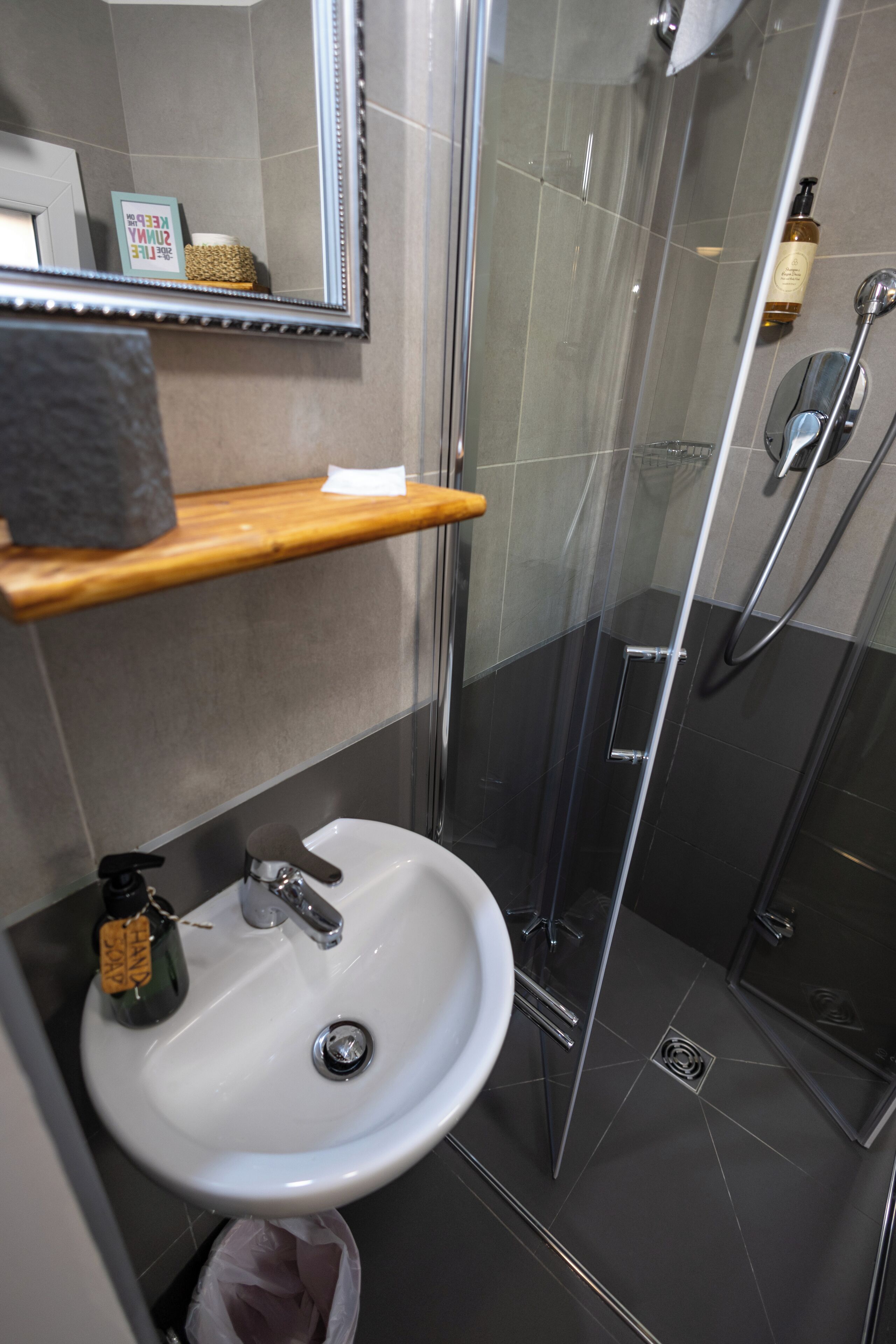 A bathroom with a white sink, a wooden shelf, a glass shower enclosure, and a mirror.