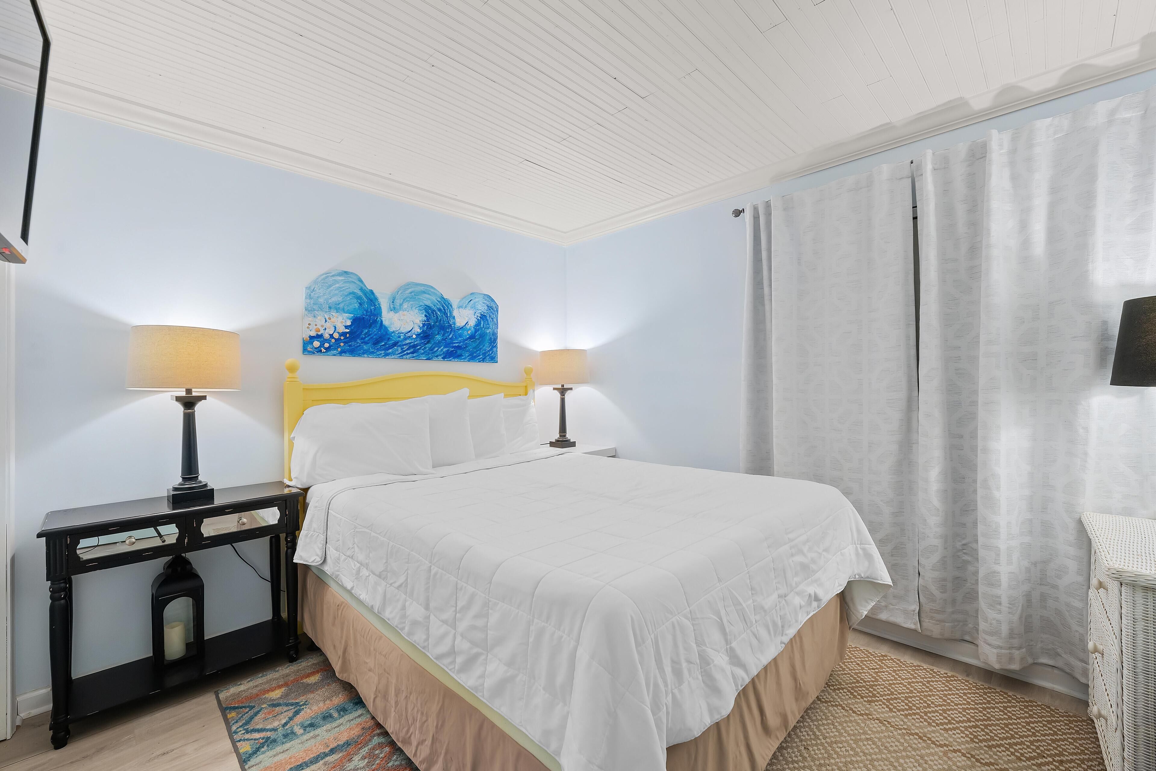 A neatly arranged bedroom with a bed, two bedside tables with lamps, a wall painting, and a window with curtains.