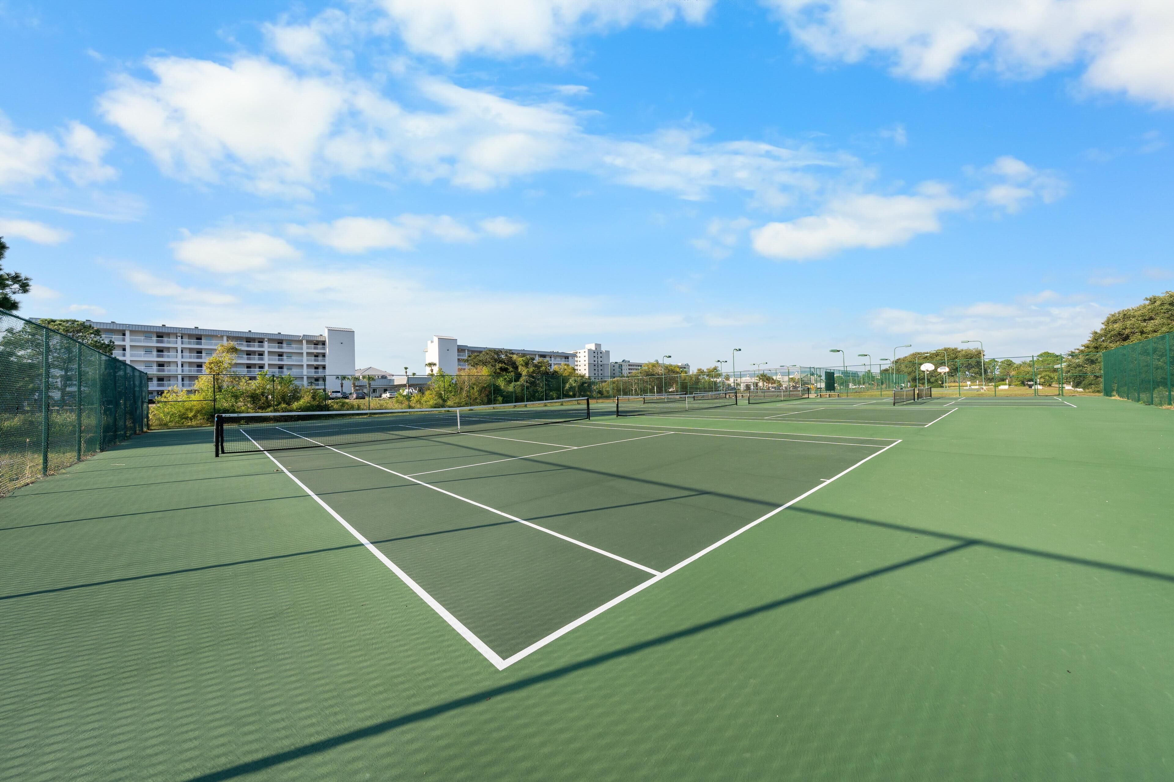 An outdoor tennis court with a green surface, surrounded by a fence, and buildings in the background.