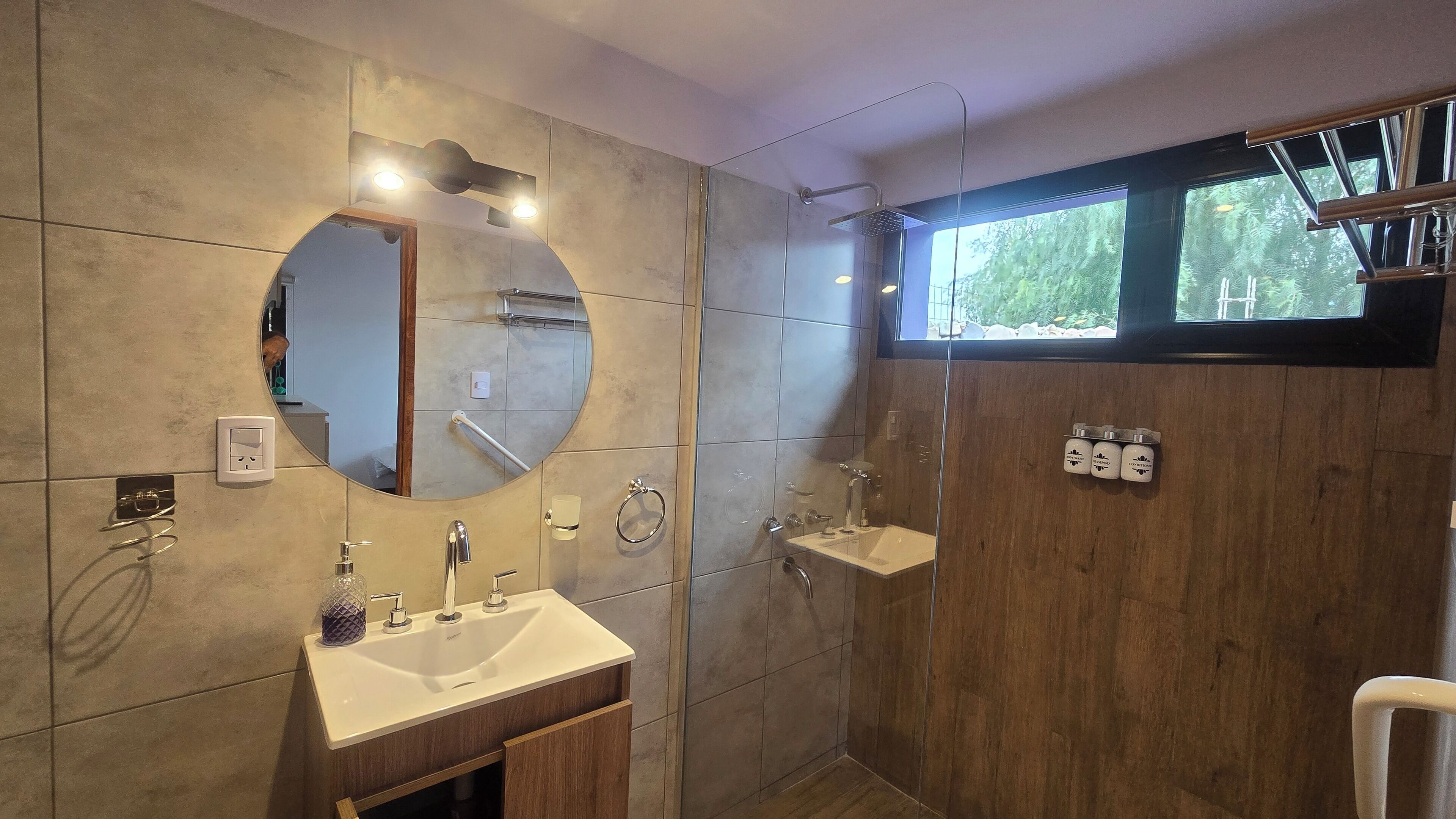 Un baño moderno con un espejo grande, un lavamanos, una ducha y una ventana con vistas a la vegetación.