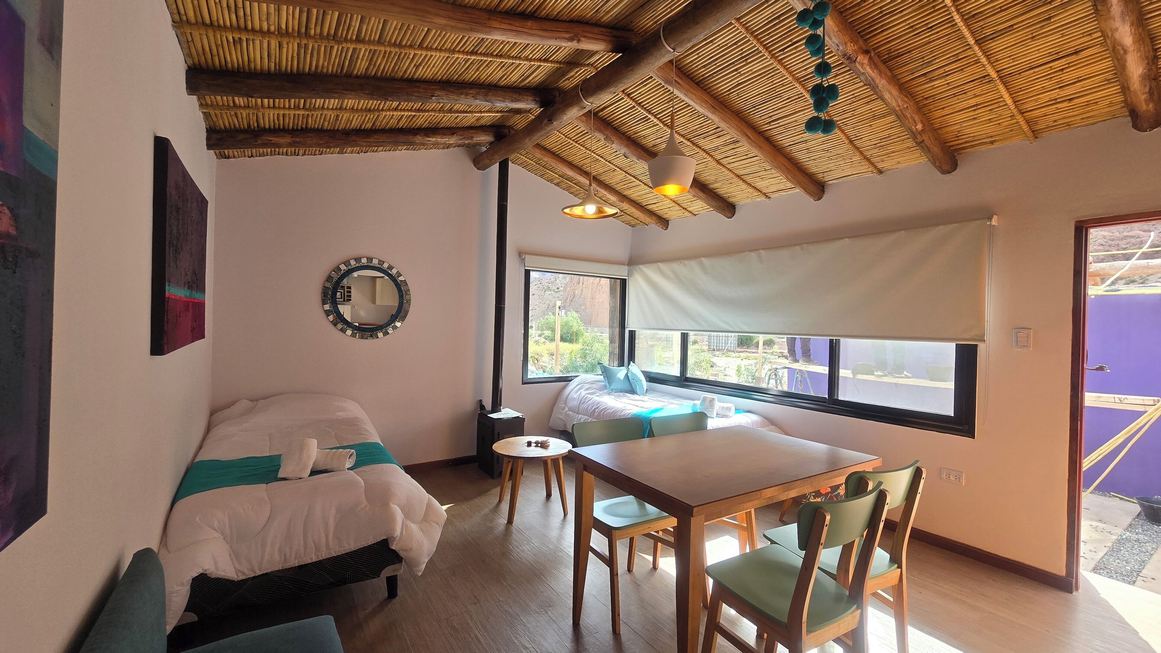 Una habitación con dos camas, una mesa de comedor con sillas, un ventanal con vistas y un techo con diseño tejido.