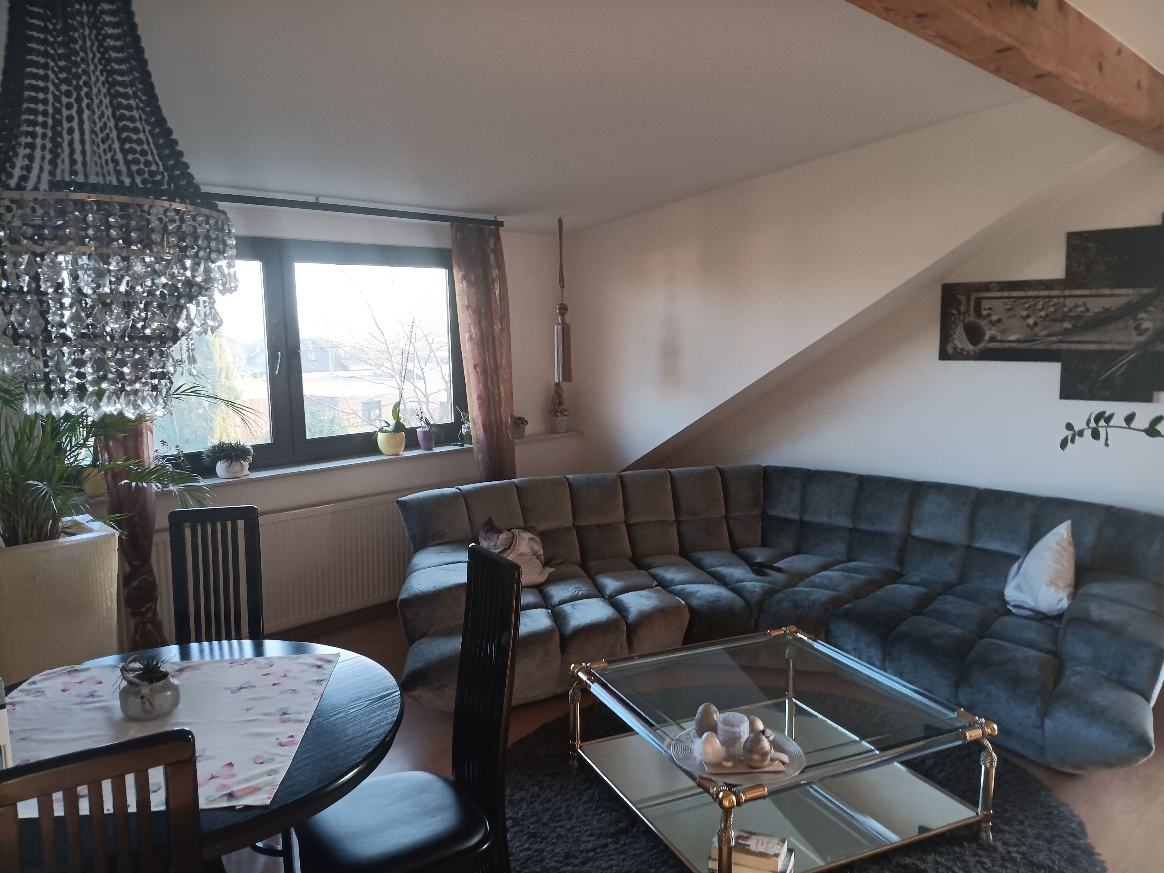 Een moderne woonkamer met een donkergrijze modulaire bank, een glazen salontafel, een eettafel met stoelen en een groot raam met gordijnen.